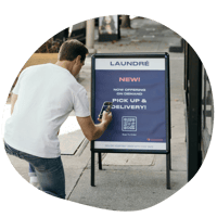 Basic Cents Design Elements-1A person scans a QR code in front of a laundromat sign that highlights a pickup and delivery service.
