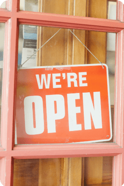 An orange sign hanging in a window that reads “We’re Open” in white font.